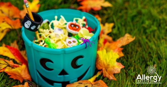 Teal plastic pumpkin filled with Halloween themed toys and surrounded by autumn leaves