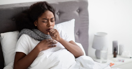 black woman in bed coughing and sneezing with medicine and tissues at her bedside table